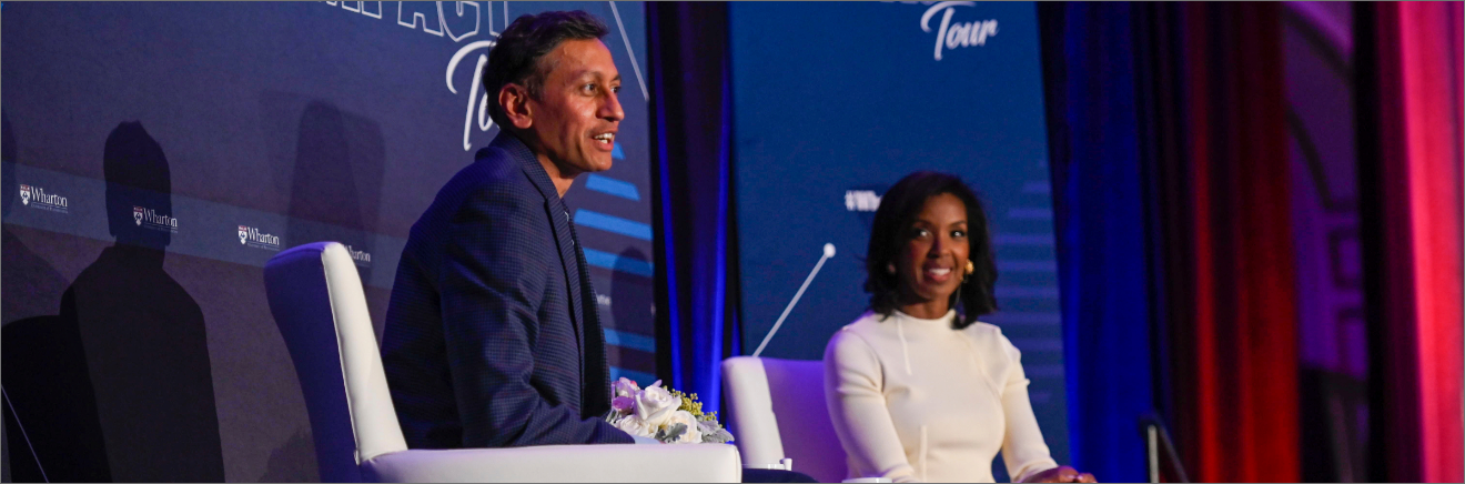 Two individuals seated on a stage during a discussion or interview event, with Wharton branding visible in the background.