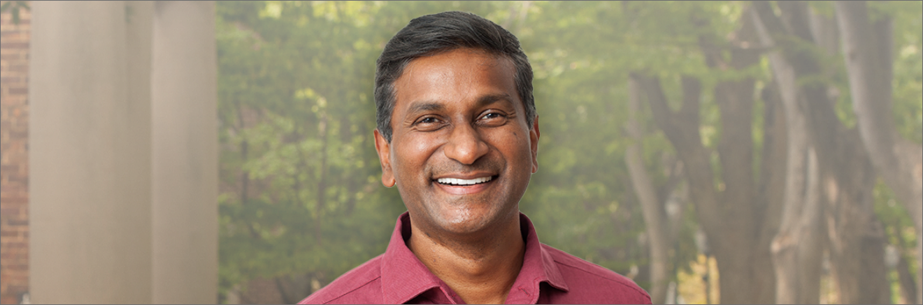 A person smiling in an outdoor setting with trees in the background. The person is wearing a red collared shirt.