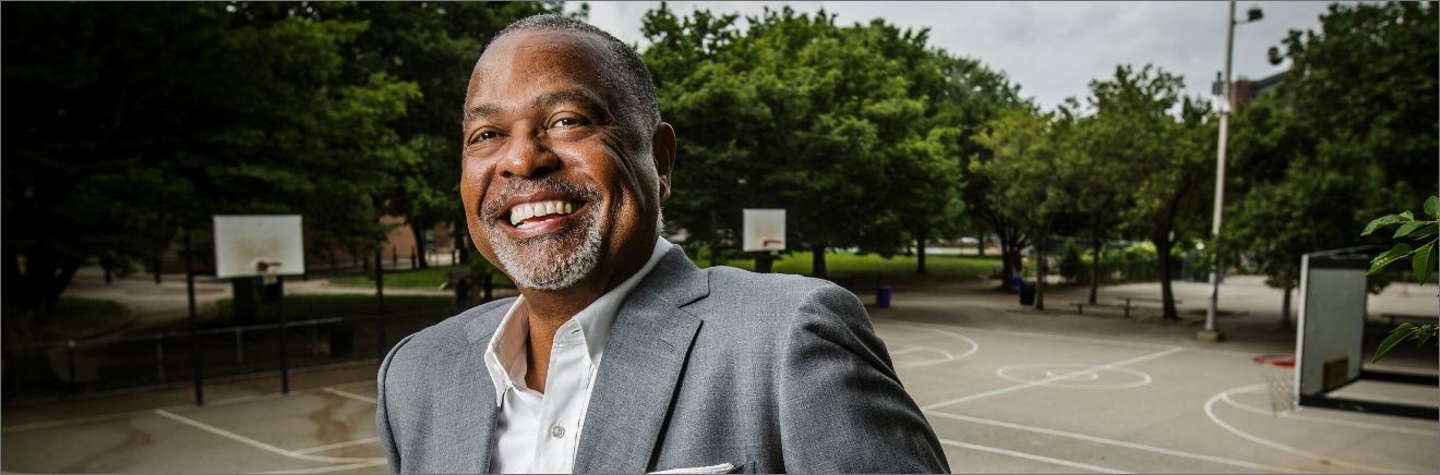 A person in a gray suit smiling in front of an outdoor basketball court with trees in the background.