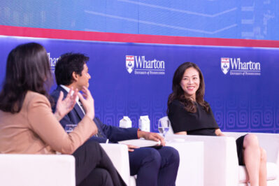 A panel discussion featuring three people seated on stage in front of a Wharton University of Pennsylvania backdrop. One person is speaking while others listen.