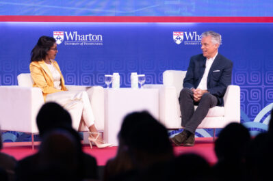 Two people engaged in a conversation on stage at a Wharton School event. They are seated on white chairs with a blue backdrop displaying the Wharton logo.