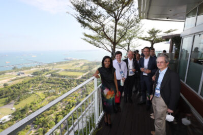 A group of people is standing together on a high balcony with a scenic view of greenery and the ocean in the background. They appear to be at a social or professional event.