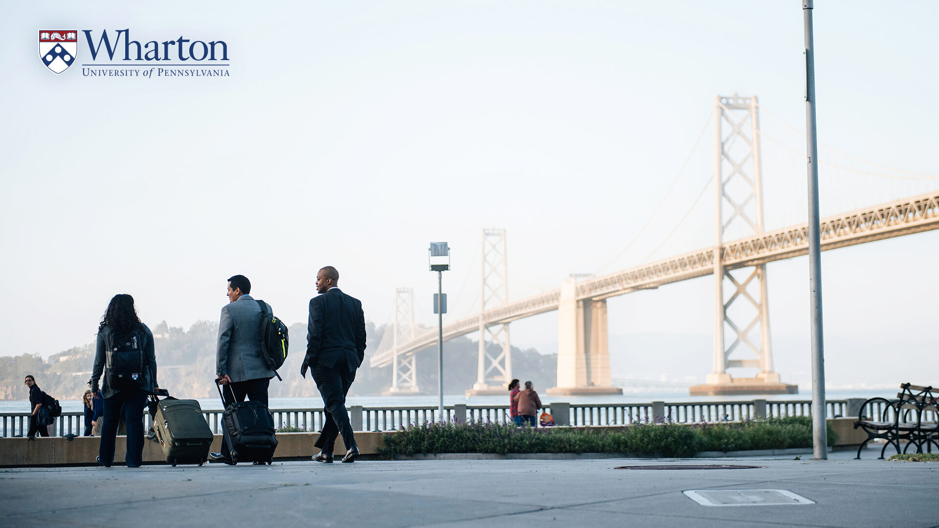 People walking on board walk near campus with a bridge in the background