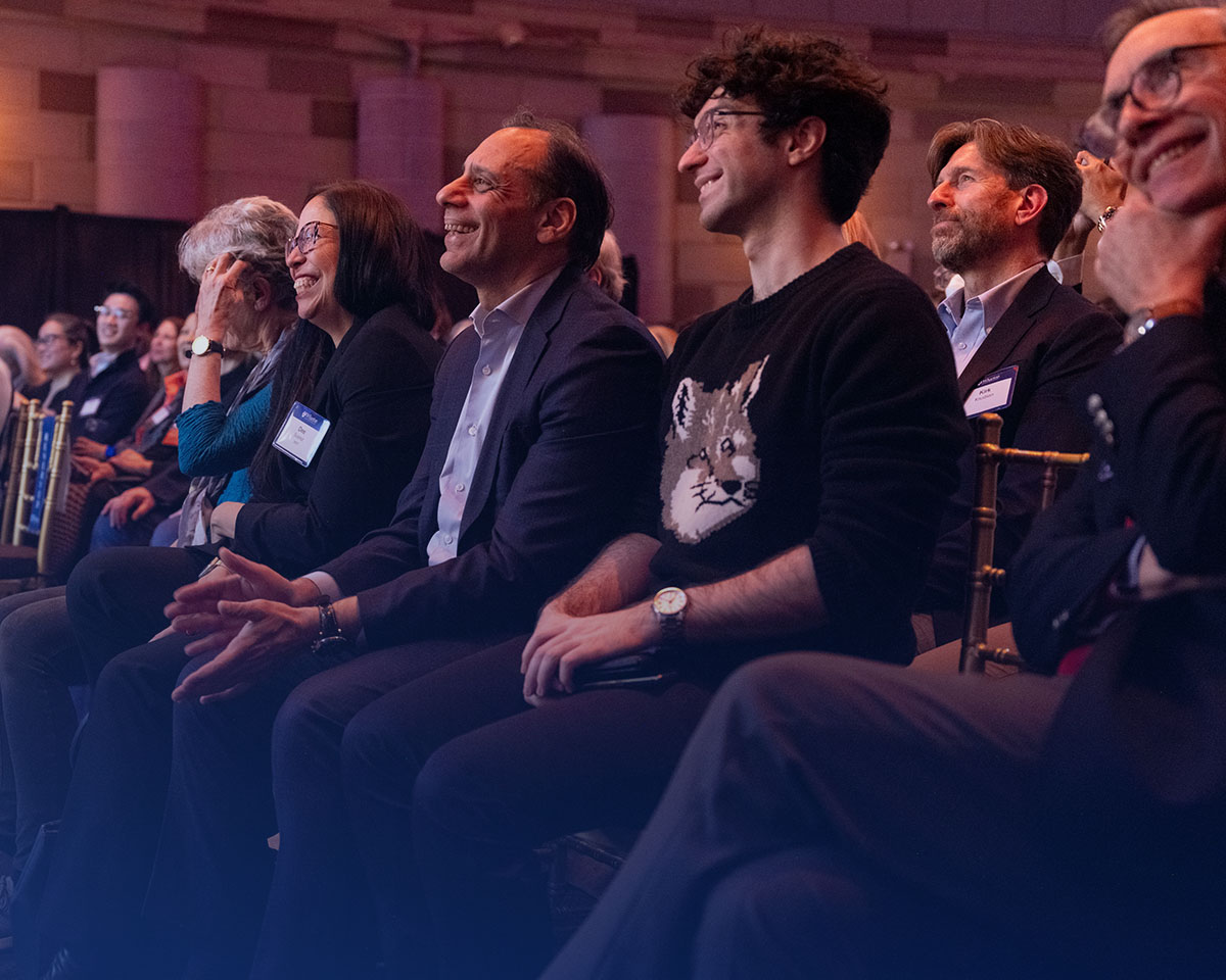 A group of people sitting in an audience, smiling and engaged, with some wearing name tags. They appear to be attending an event or presentation.