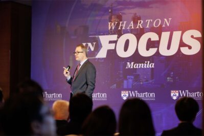 A person in a suit speaks at a Wharton event in Manila. The backdrop features the words 