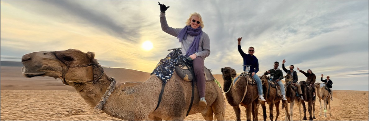 A group of Lauder Institute students riding camels in a desert with cloudy skies and the sun partially visible.