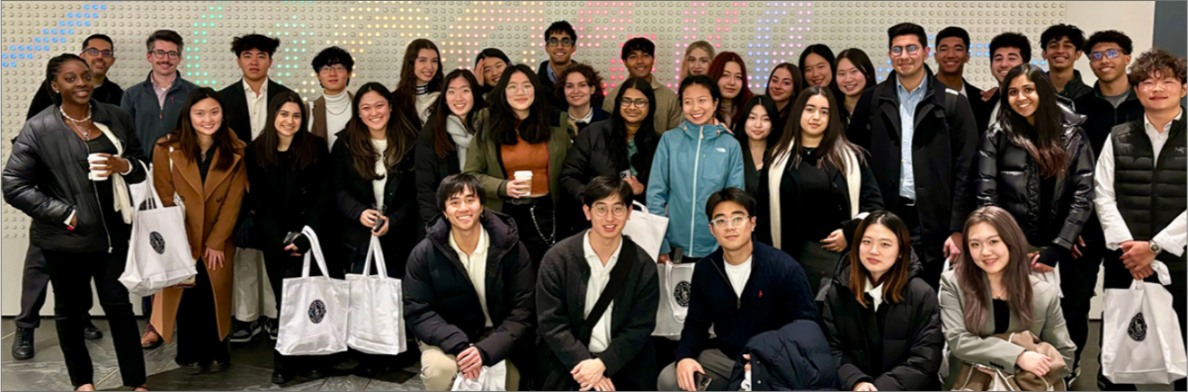 A large group of diverse Wharton students posing in winter attire in front of a lit perforated wall.