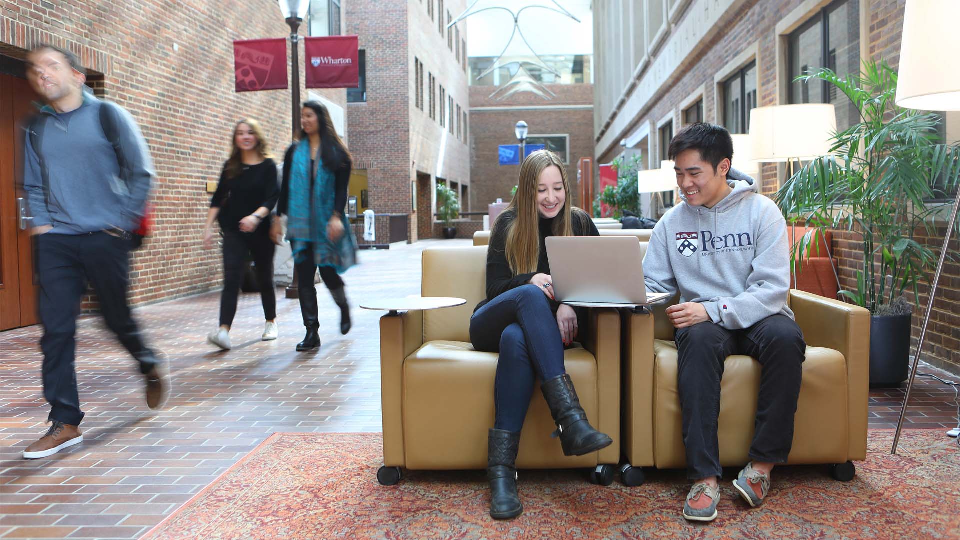 Wharton Online Course - Header 2 Two Wharton alumni sitting and smiling at a laptop in a brick-walled university corridor.