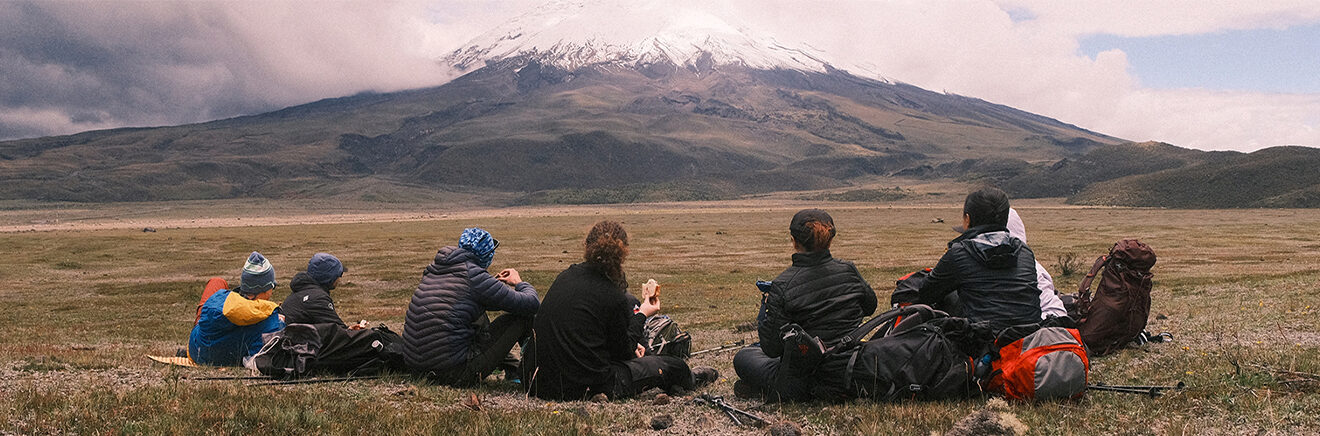 A group of Wharton students sitting in a field in Ecuador, observing a snow-capped mountain in the distance.