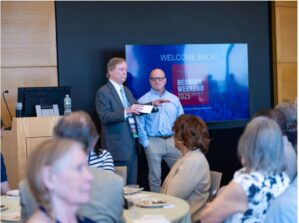 A person in a suit and another in a shirt are speaking in front of an audience at a reunion event. The screen behind them reads 