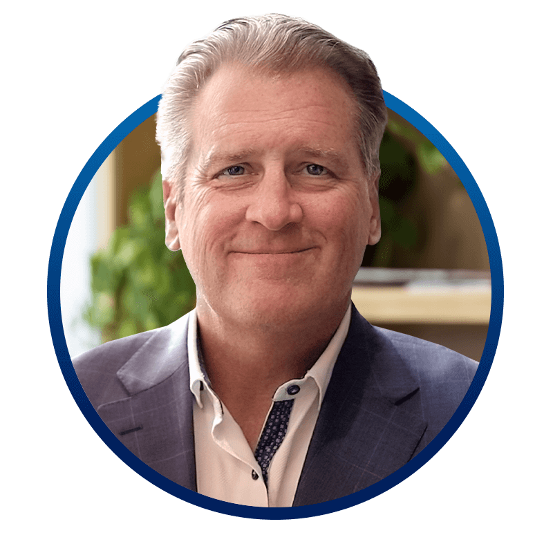 Headshot of a smiling person with light-colored hair, wearing a suit and collared shirt, set against a blurred indoor backdrop with greenery.