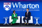 A person in a green suit and another in a dark suit sit on a stage with "Wharton University of Pennsylvania" in the background.