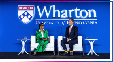 A person in a green suit and another in a dark suit sit on a stage with "Wharton University of Pennsylvania" in the background.