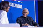 A couple of people are seated and engaged in conversation on a stage. One person is smiling, and the backdrop displays the word "PENNSYLVANIA."
