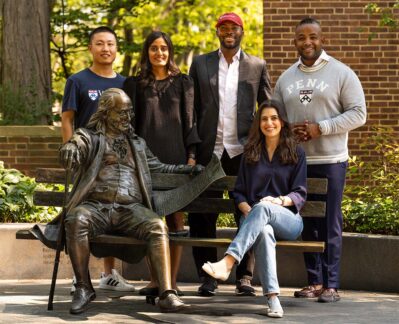 Five Wharton fellows pose around a bronze statue of Benjamin Franklin on a bench.